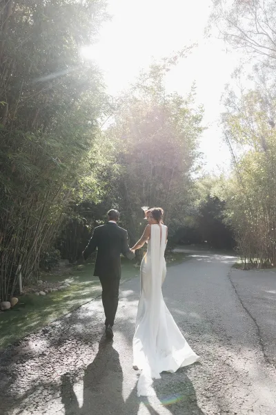 Couple portrait of the bride and groom walking hand in hand along a paved path, her long veil trailing, framed by a tranquil bamboo grove.