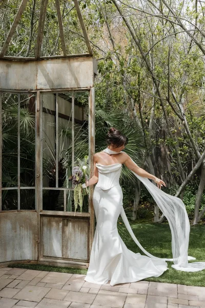 Bridal portrait of a bride with bouquet of calla lilies, standing under garden trees by a rustic metal and glass structure on stone pavers.