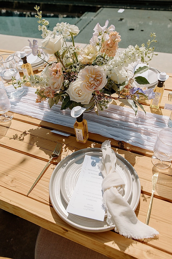 Reception tablescape with wedding table centerpiece of white and blush roses, greenery, gold flatware and glassware on a sunlit outdoor wood table