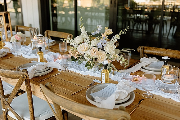 Reception tablescape on a wood farm table wedding with gauze runner, rose centerpiece, candles, gold flatware, and place cards by glass doors