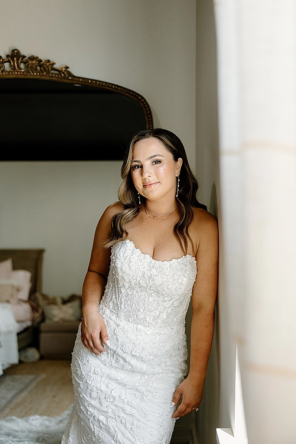 Bridal portrait of a bride in a strapless lace wedding dress, leaning by a bed in soft window light, wearing drop earrings and ring