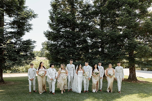 Wedding party portrait with bride and groom and bridal party holding white and greenery bouquets on a lawn with evergreen trees backdrop