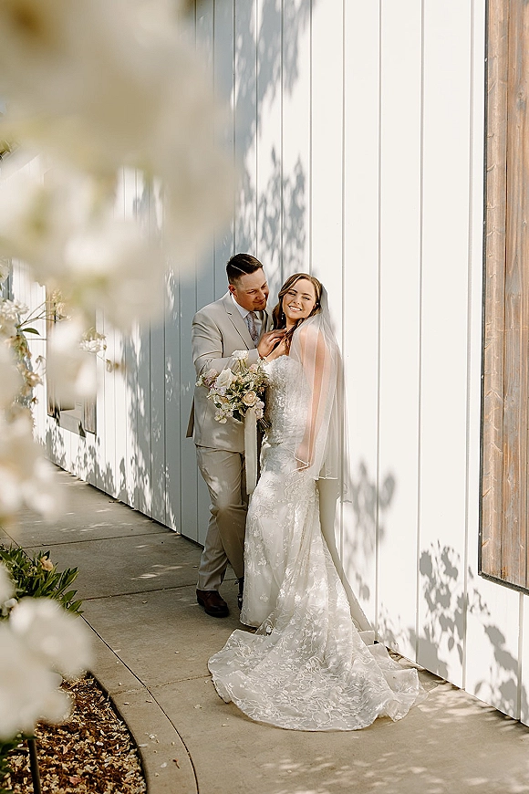 Couple portrait of bride and groom posing as he whispers to her, bouquet and veil visible against a white paneled wall in dappled light