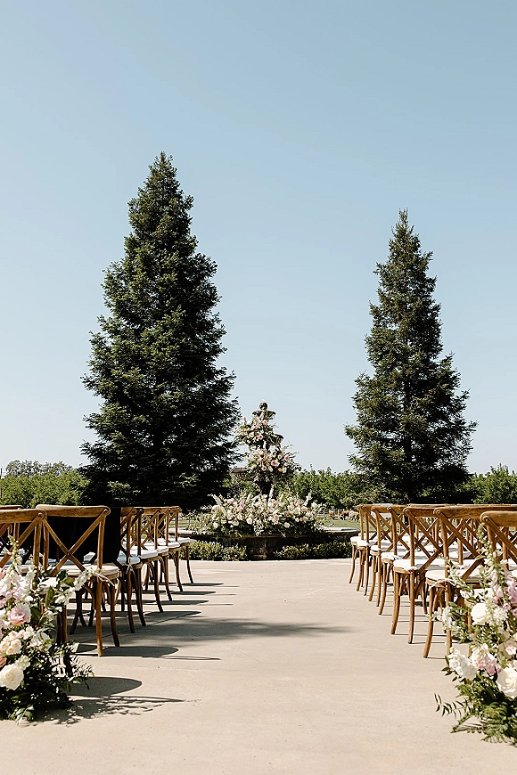 Ceremony setup for an outdoor wedding ceremony with wood cross back chairs lining a paved aisle, pink and white flowers by a fountain under blue sky
