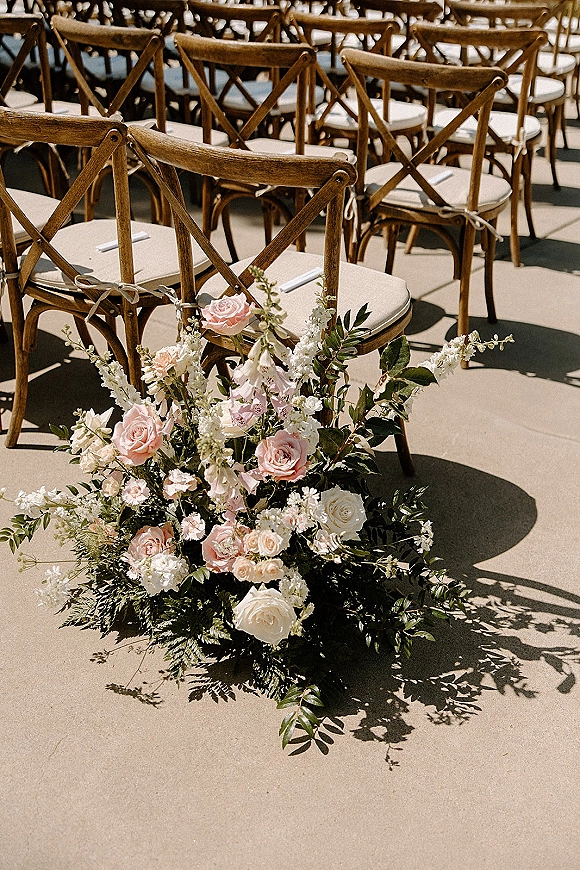 Ceremony aisle decor with an aisle floral arrangement of white roses and greenery beside cross back chairs on a sunlit concrete floor