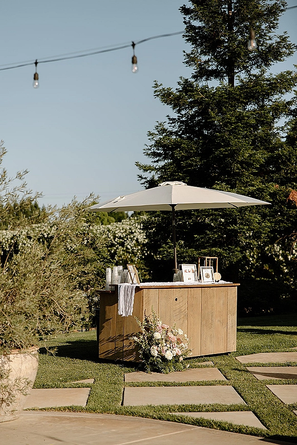 Outdoor bar setup with a wood bar under a patio umbrella, string lights and framed signs, plus blush floral arrangement on lawn pavers