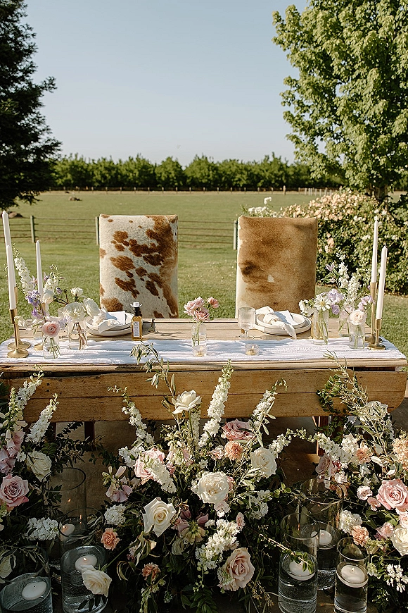Sweetheart table decor with an outdoor sweetheart table setup, taper candles and brass candlesticks among roses and greenery in a pasture field backdrop