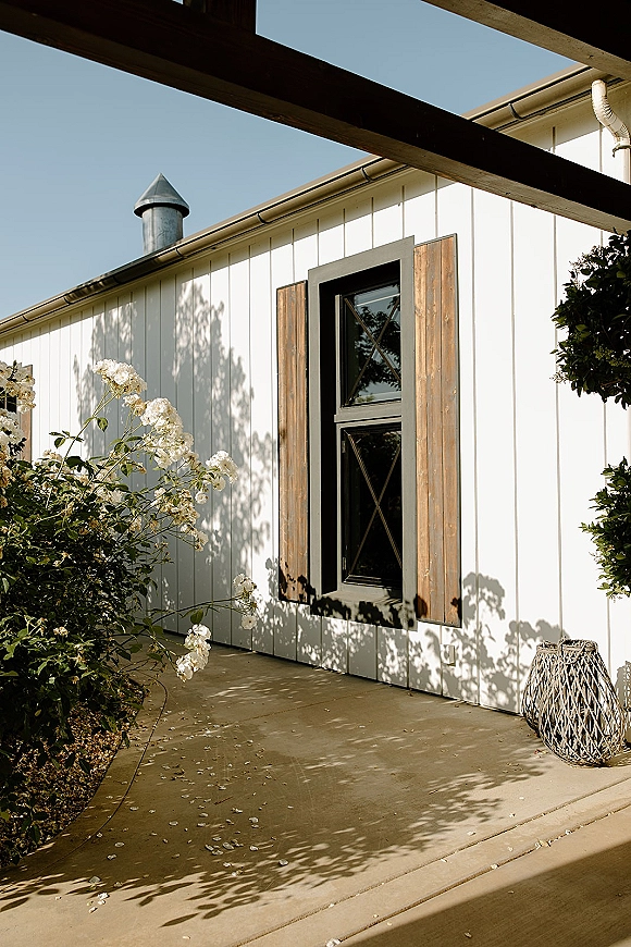 Outdoor venue exterior with a black-framed window, wooden shutters, and white flowers with greenery beneath pergola beams on a patio