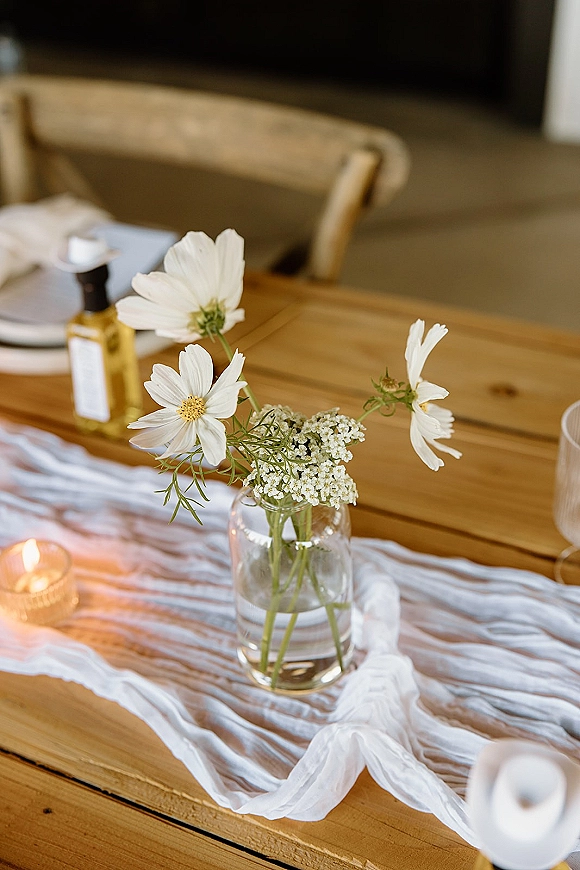 Reception table decor with bud vase centerpiece of white flowers, gauze runner, votive candle, and place setting on a wood farmhouse table