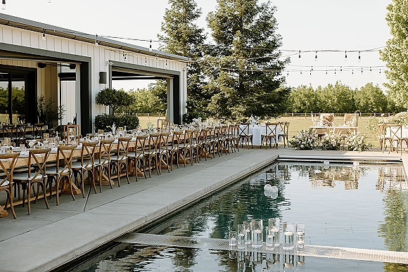Outdoor reception setup with long banquet tables dressed in white linens, candles and floral centerpieces, beside a pool at a modern barn patio