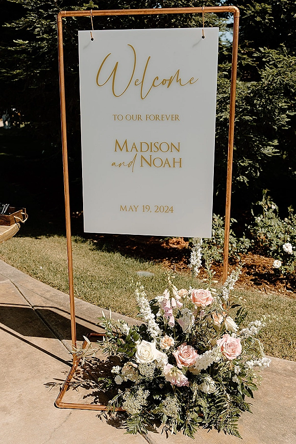 Wedding welcome sign with flowers in gold calligraphy on a white board, set on a copper stand with blush roses and greenery in a garden walkway