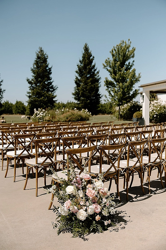 Ceremony seating with outdoor wedding chairs in neat rows, white cushions and rose-and-greenery aisle florals on a sunny lawn near trees