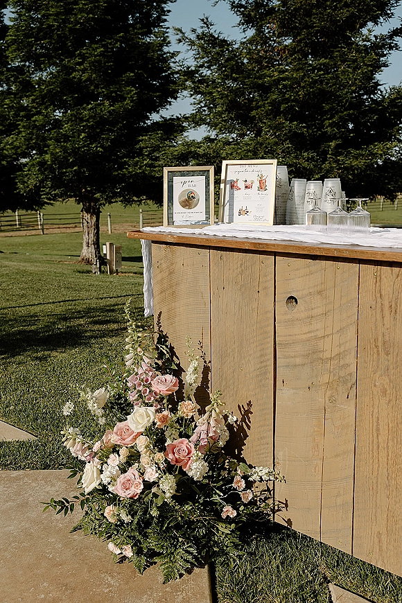 Wedding bar setup with framed signage, menu sign, and glassware on a rustic wooden bar, finished with rose and greenery accents on a lawn