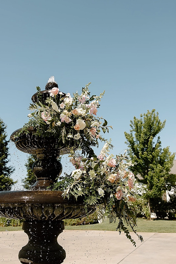 Wedding fountain decor with a fountain floral arrangement of pink roses, white blooms, and trailing greenery around a stone fountain spray under blue sky