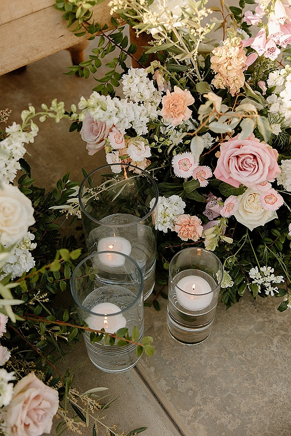 Wedding floral decor with blush and white roses and greenery in glass cylinder vases, floating candles glowing beside a wooden bench on stone floor