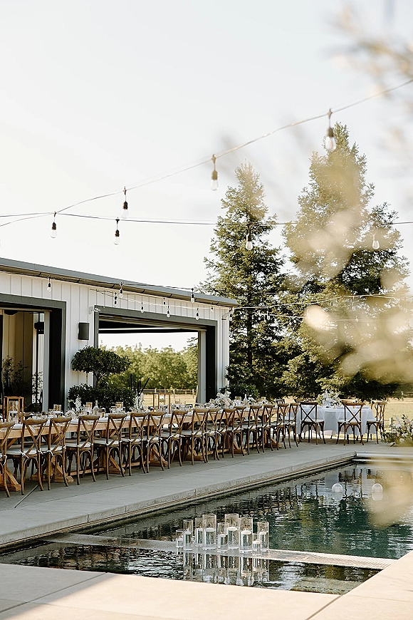Outdoor reception decor with poolside wedding reception setup, long banquet table in white linens, crossback chairs, string lights by modern barn patio