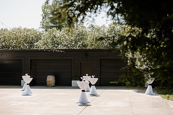 Cocktail hour setup with wedding cocktail tables in white linens and black sashes, floral centerpieces under string lights by a black building exterior