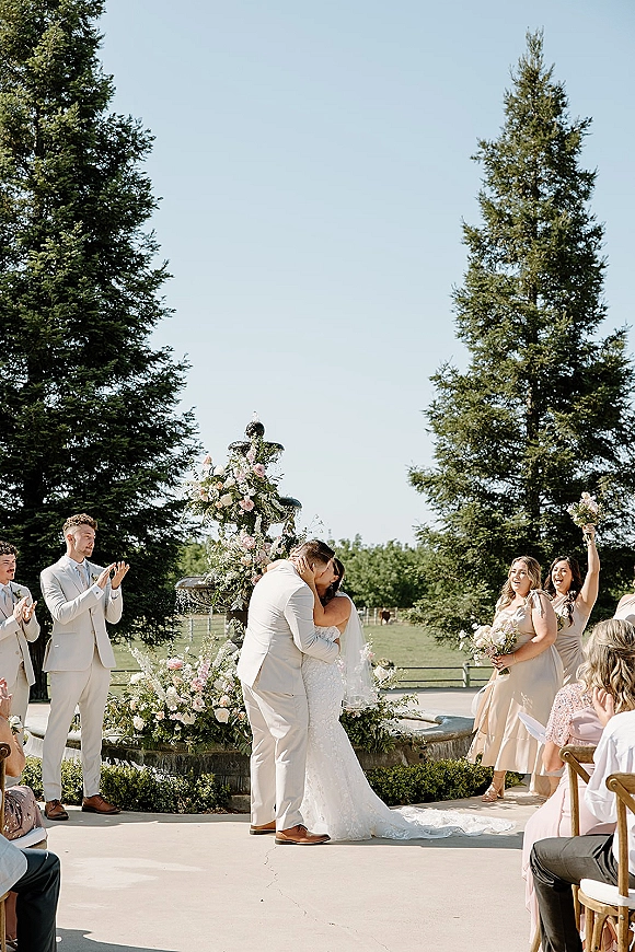 Wedding kiss portrait of bride and groom kissing in front of a fountain, with bridesmaids cheering and guests seated on a sunny lawn