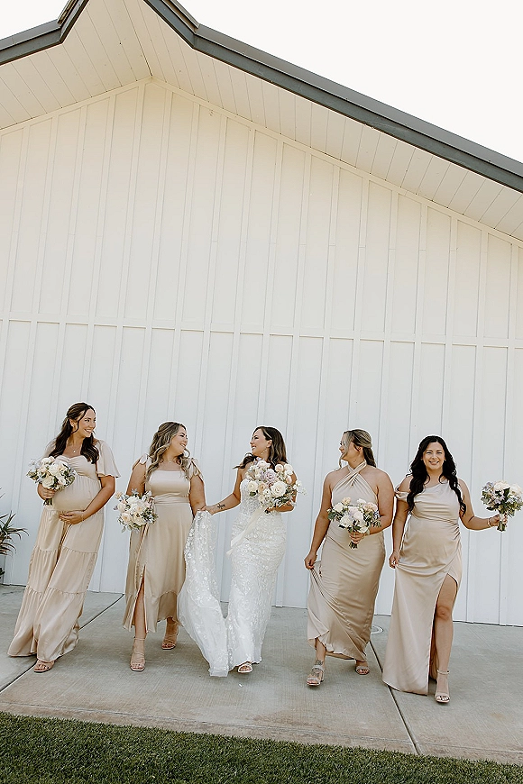 Bridesmaid group photo with bride and bridesmaids walking in neutral dresses, holding white bouquets by a white barn wall