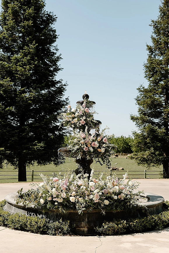 Wedding fountain decor with pink and white rose florals cascading over a tiered stone fountain, set in a paved courtyard with evergreens