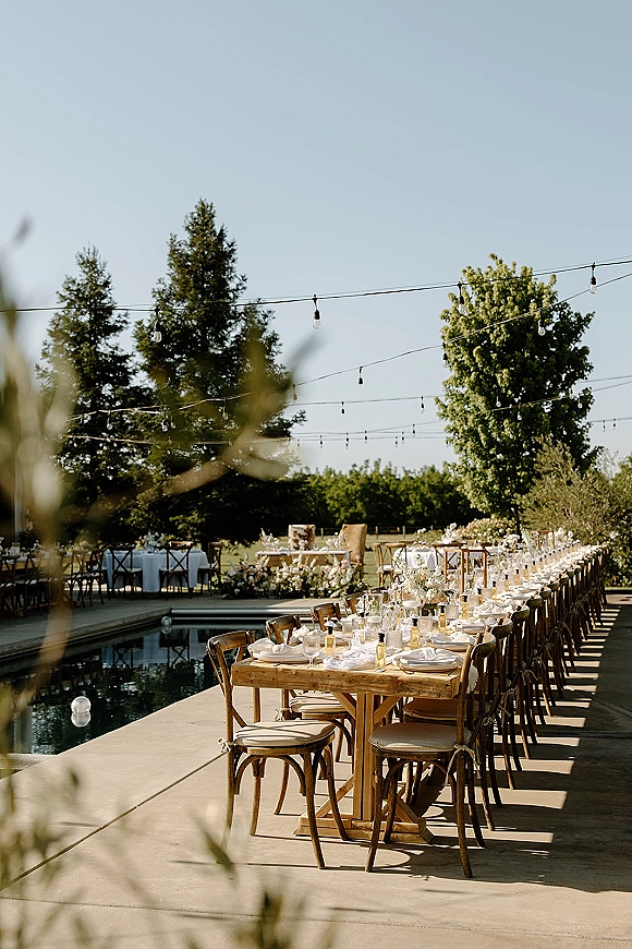 Reception tablescape with a long wedding reception table set with white napkins, candles, floral garlands and string lights by the pool outdoors