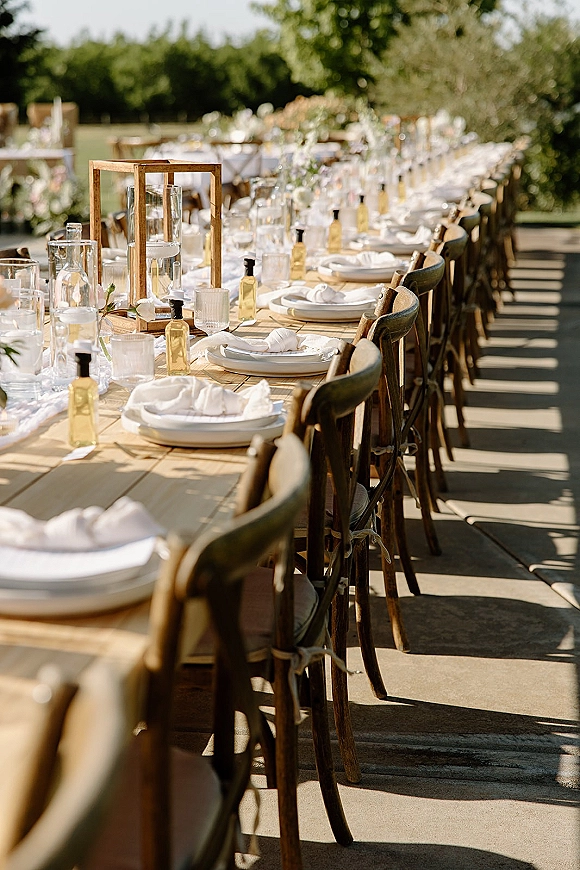 Reception tablescape with an outdoor wedding reception table on a lawn, set with white plates, napkins, glassware, lanterns and greenery runner