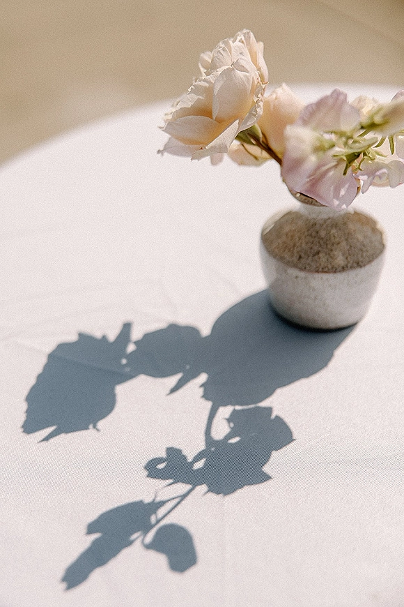 Bud vase centerpiece with a blush flower and green stems in a small stone vase on a white tablecloth in hard sunlight with floral shadows