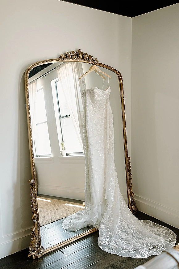 Wedding dress, lace wedding gown hanging on a wooden hanger, reflected in a vintage gold mirror by sheer curtains and window light