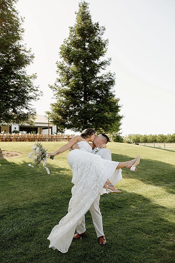 Wedding kiss portrait of groom lifting bride in a dip, her lace strapless gown and bouquet framed by a green lawn and evergreen tree.