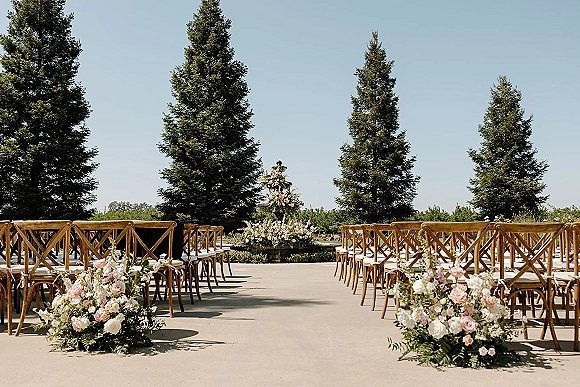 Ceremony setup for an outdoor wedding ceremony with cross back chairs, white cushions, and blush floral aisle arrangements in a vineyard lawn setting