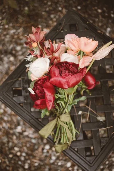 Bridal bouquet of red peonies and ranunculus with greenery and ribbon wrap resting on a metal patio table over gravel ground