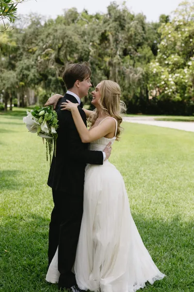 Couple portrait of bride and groom embrace, her white calla lily bouquet visible on a daylight garden path beneath mossy trees