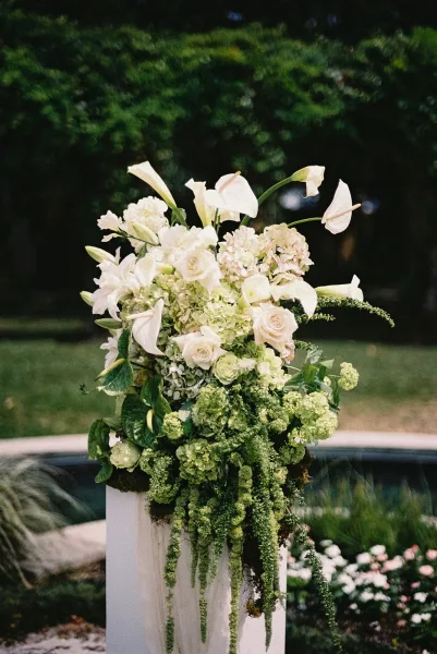 Wedding ceremony flowers cascade from a floral pedestal with white calla lilies and hanging amaranthus on a garden lawn pathway