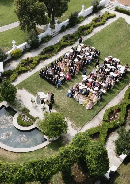 Outdoor wedding ceremony seen from above with white parasols and chair rows on a manicured lawn beside a pond fountain