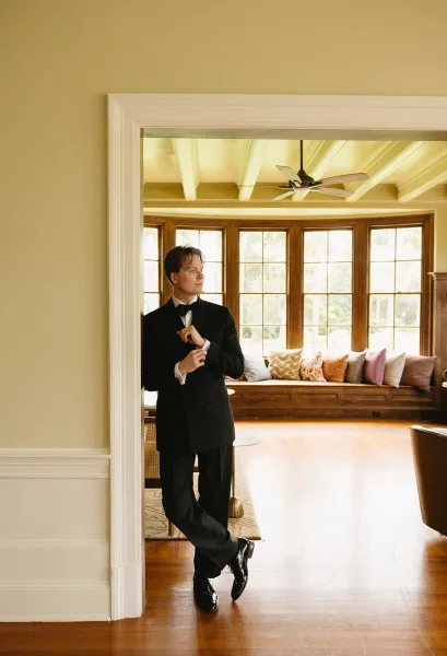 Groom portrait in a classic black tuxedo, adjusting cufflinks in an interior doorway with wood floors and bay windows behind him