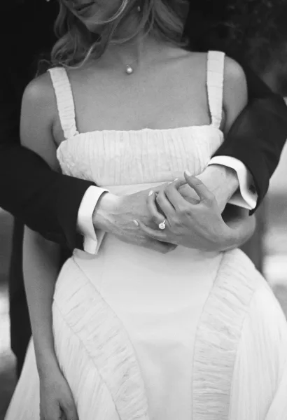 Couple portrait of bride and groom embrace, groom hugging bride as her diamond ring rests on his hand against a dark backdrop
