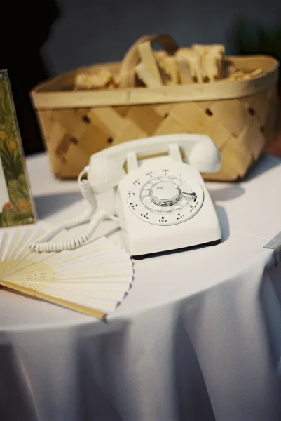Wedding guest book phone setup with a white rotary telephone beside a wicker basket and paper hand fan on a white tablecloth indoors