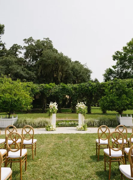 Ceremony setup with floral arrangements lining a gravel aisle, wooden chairs with white cushions, and a greenery hedge arch on a garden lawn