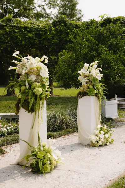 Ceremony entrance decor with wedding aisle entrance floral pillars, white draping, roses, orchids, and greenery along a gravel garden path