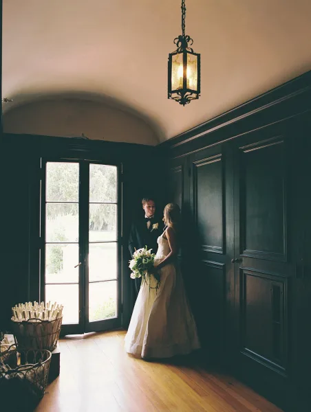Couple portrait of bride and groom portrait by a tall window, bride in strapless dress holding bouquet, groom in black tuxedo in wood-paneled room