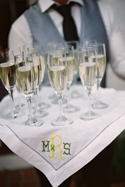 Champagne toast with sparkling wine in flutes beside an embroidered monogram cocktail napkin, set on a table near a groom’s suit and tie
