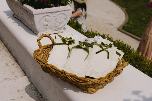 Wedding programs in a wicker basket tied with green ribbon bows on a white ledge beside a stone planter and greenery outdoors
