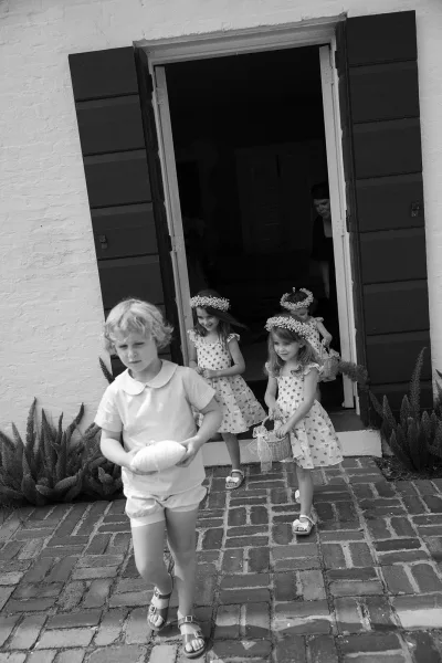 Wedding flower girls in matching dresses with flower crowns, holding a basket and ring pillow on a brick walkway by an open doorway