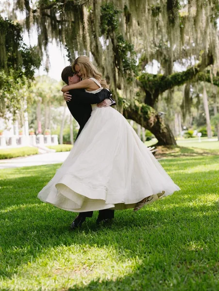 Wedding kiss portrait of groom lifting the bride as they kiss, her tulle dress swirling beneath an oak tree draped in moss in daylight
