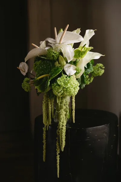 Bridal bouquet with calla lily bouquet styling, featuring white calla lilies and anthurium with trailing amaranthus in a glass vase against a dark wall