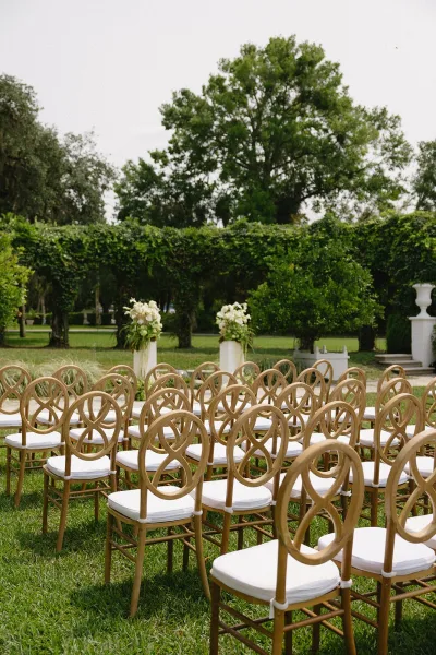Ceremony seating with wood chairs and white cushions lined along a white aisle runner on garden lawn, framed by floral pedestal columns
