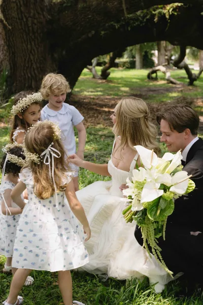 Couple with flower girls seated on park lawn as the bride chats with them, holding a calla lily bouquet under dappled tree light