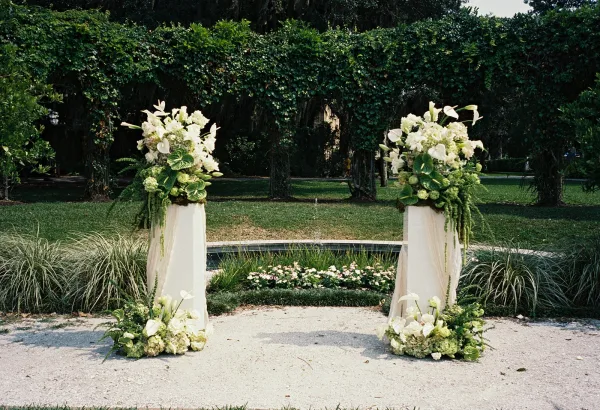 Wedding ceremony arch with white and green wedding flowers, draped fabric and calla lilies on pedestals in a garden near a fountain