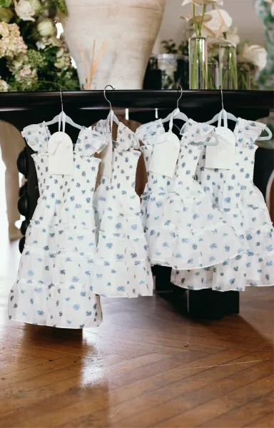 Flower girl dresses in blue floral and white hang from tagged hangers, featuring ruffled straps and tiered skirts on a wood floor by a grand piano