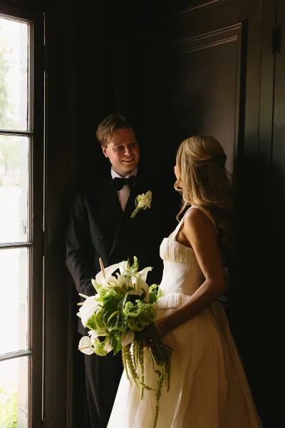 Couple portrait of bride and groom indoors, her strapless gown and calla lily bouquet beside his tuxedo in soft window light